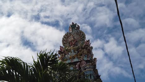 a shot of the tower of a hindu temple full of deities as walking down the street, with a palm in the middleground and many tiny clouds in the sky