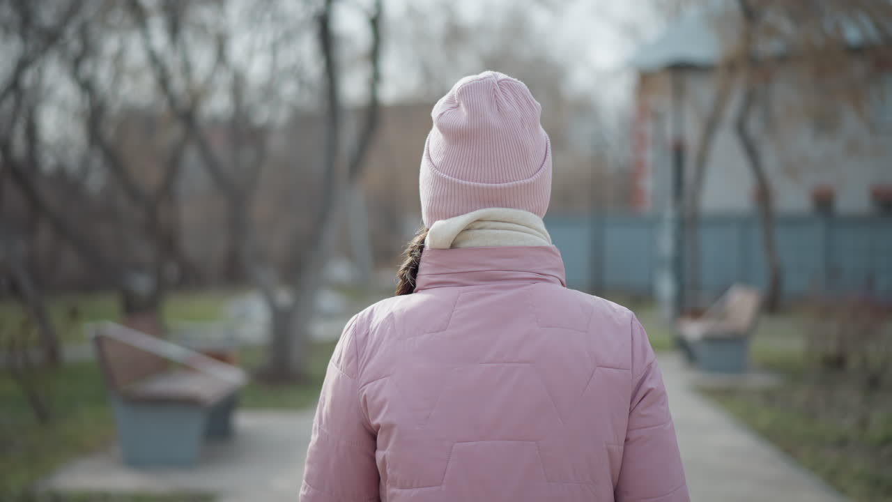 Woman in pink winter jacket and knitted pink hat viewed from behind, standing still in quiet park during cold day, with soft background of trees, benches, and blurred urban environment around
