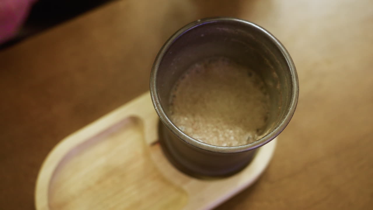Primo piano della mano di una signora con una giacca di montone marrone che solleva delicatamente una tazza di caffè a un accogliente tavolo da bar. L'illuminazione soffusa crea un'atmosfera invitante e calda, perfetta per momenti di pace.