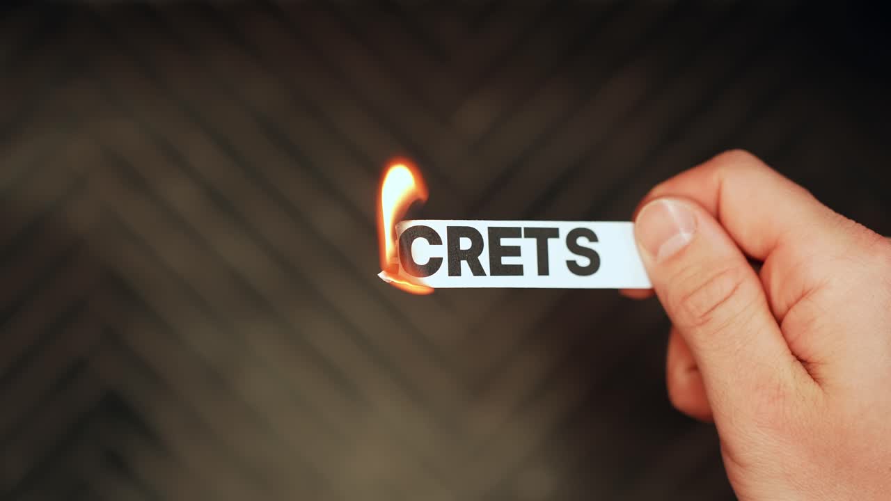 Primo piano di una mano che tiene una striscia di carta recante la scritta “SECRETS” mentre una fiamma ne brucia il bordo, simboleggiando verità nascosta, confessione, rivelazione e la rottura del silenzio.