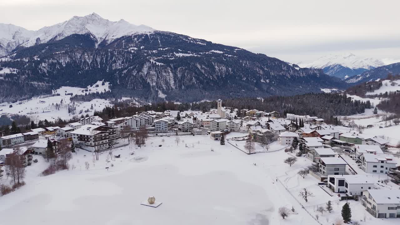 Vista aérea de Laax, Suiza, que muestra un pueblo alpino cubierto de nieve con una iglesia central, campos helados y los Alpes circundantes. Ambiente invernal tranquilo bajo un cielo encapotado en Europa. Plano general