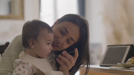 Happy-Beautiful-Woman-Sitting-At-Kitchen-Table-And-Feeding-Her-Baby-Daughter-With-Fruit-Puree