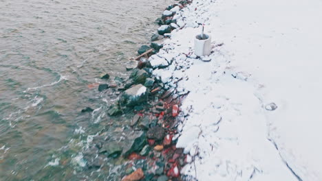 Flying-over-large-black-and-red-rocks-on-a-winter-beach-with-water-and-waves-moving-on-other-side,-while-ground-is-covered-in-snow