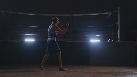 professional beautiful female boxer shoots off conducting a shadow fight in a dark hall room in slow motion in blue clothes and red bandages on her wrists. steadicam shot