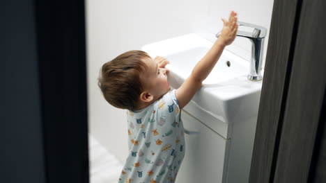 curious smiling toddler standing in the bathroom. cute baby boy playing with water faucet. top view.