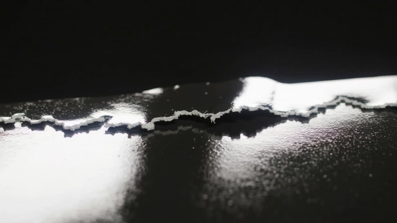 Strappo di una pellicola nera lucida che espone un substrato bianco sotto la luce dello studio, con la formazione di un lembo e il sollevamento di scaglie.