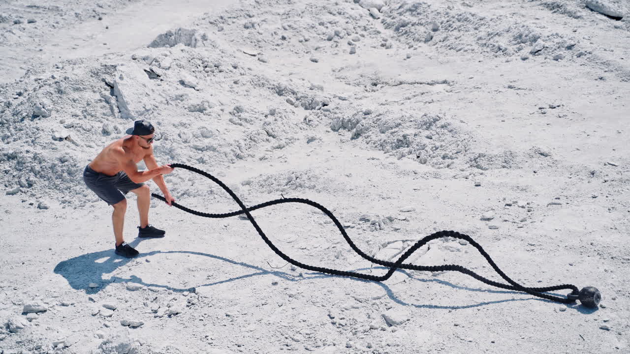 Athletic male performs battle ropes exercise on the white rocky background. Sportsman in shorts doing waves with cable during an outdoor fitness workout.