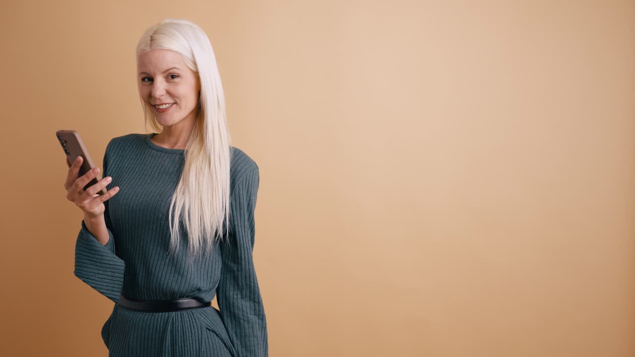 Mujer rubia feliz sosteniendo un smartphone y señalando un espacio para texto