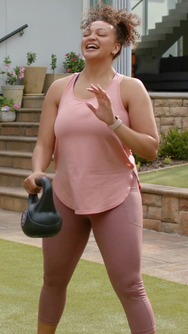 Video verticale: Una donna in capri rosa si china e solleva un kettlebell da 5 kg sul prato del patio, sorridendo.
