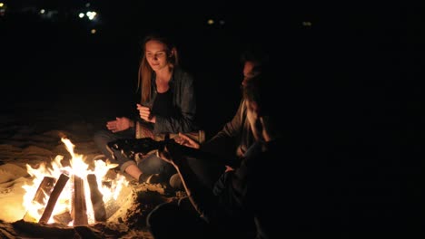 movimiento de cámara redonda: grupo multirracial de jóvenes sentados junto a la hoguera tarde en la noche y cantando canciones, tocando la guitarra y la percusión