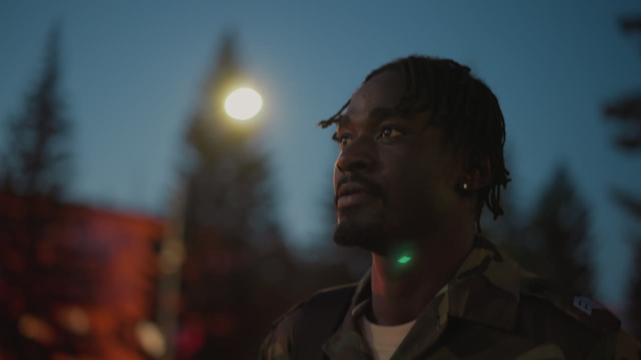 Black Soldier Under Streetlight At Dusk, Wearing Camouflage Jacket And Backpack, Pine Trees And Warm Bokeh Lights In Background, Closeup Portrait With Contemplative Expression, Cinematic Shallow