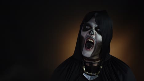 Creepy man with skeleton makeup in hood making angry faces, looking at camera, trying to scare