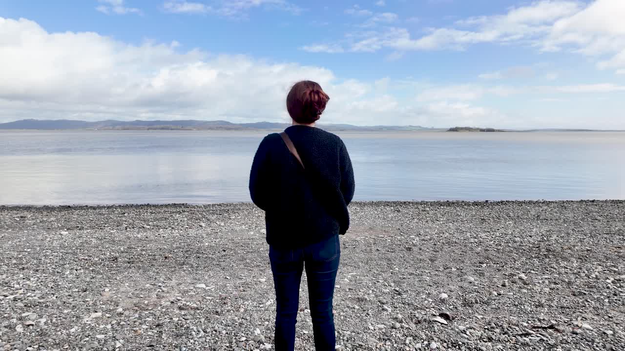 Mulher em pé em praia de seixos, observando a água calma e o horizonte, encontrando solitude.