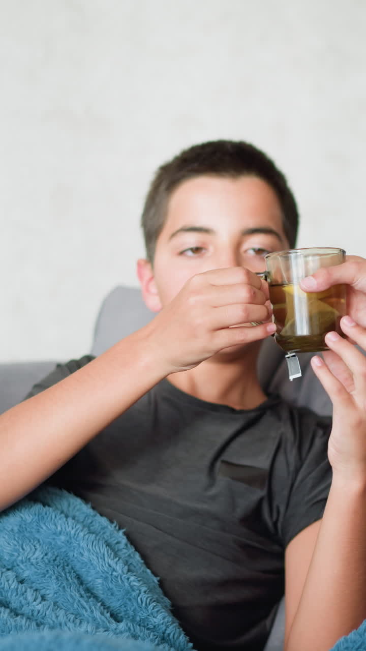 Sick boy resting on couch collects tea cup from his sister and drinks it as part of recovery process, with a concerned and caring sibling offering support