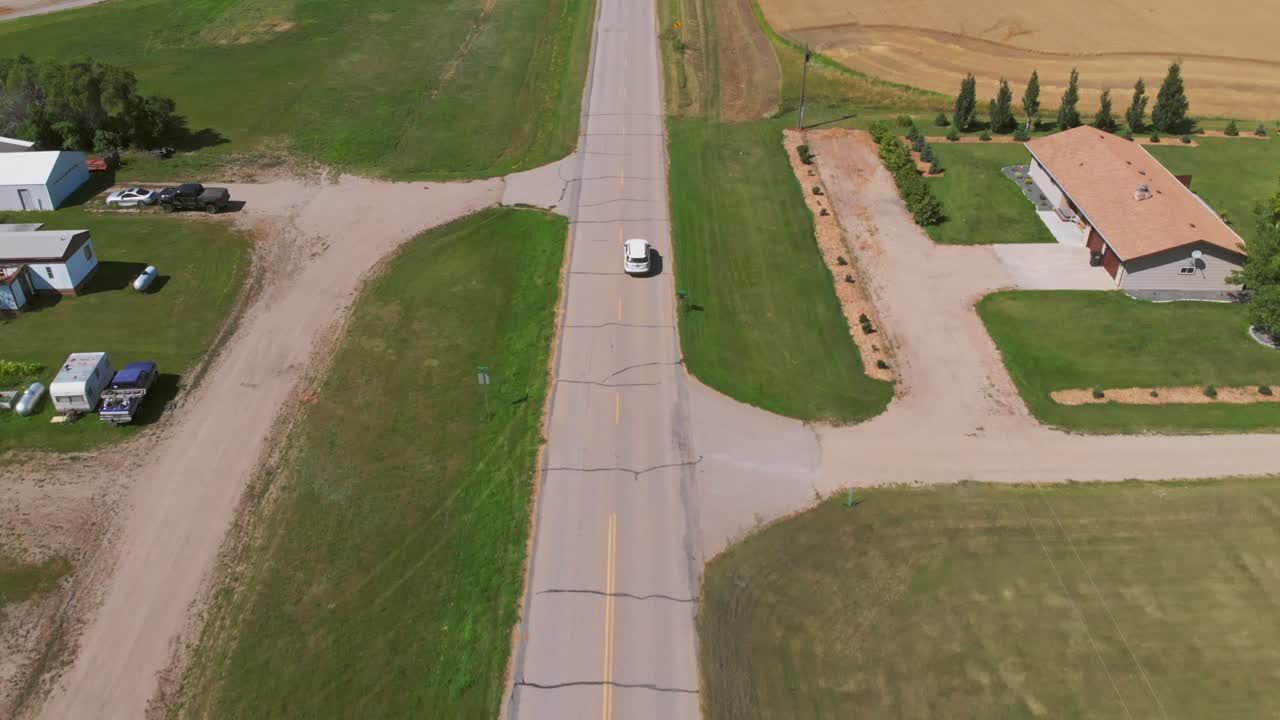 Aerial tracking shot at a slight downward angle of a white car driving on a rural road bordered by green grass and trees