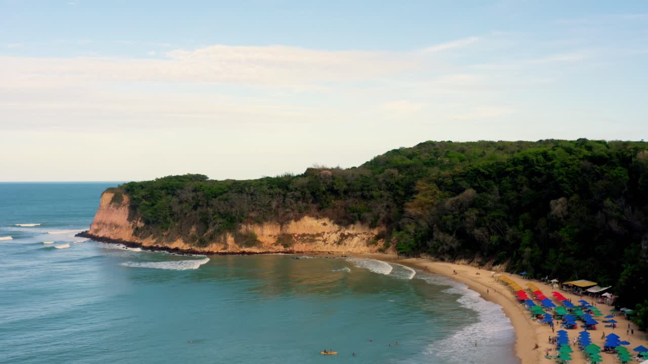 Spettacolare e ampia ripresa aerea in ascesa con drone della spiaggia tropicale Praia do Madeiro con ombrelloni colorati, che rivela la costa sopra le scogliere vicino alla famosa città di Pipa nel Brasile settentrionale.