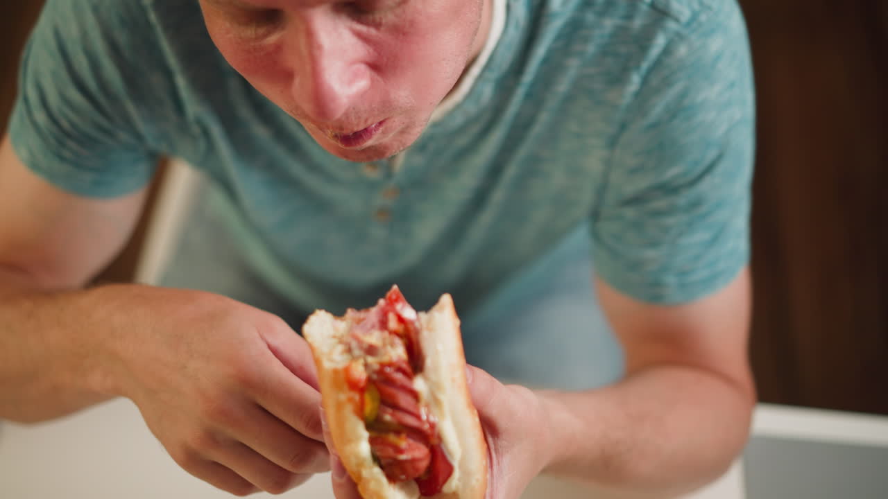 Close up of hungry boy biting into juicy burger for dinner, holding sandwich with both hands over white table, wearing casual teal shirt, filled with sausage, tomato, sauce and peper