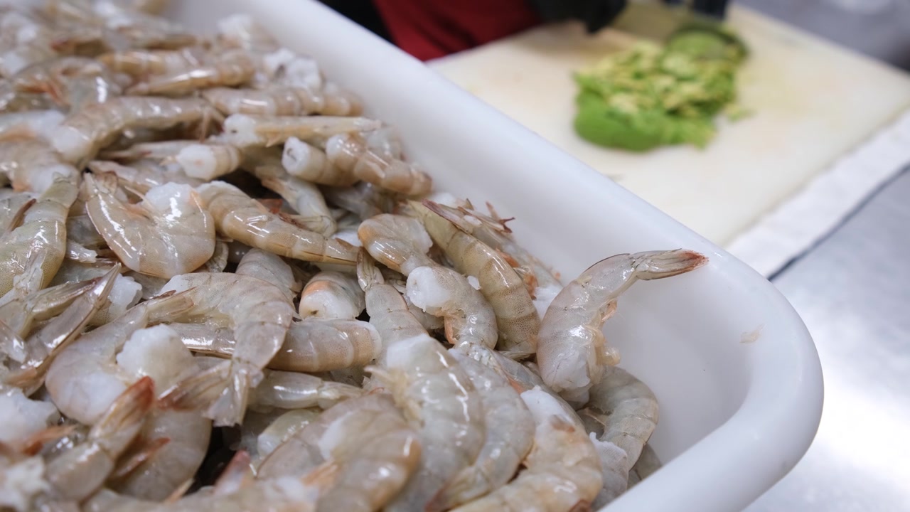 Primo piano di gamberi freschi crudi pronti per la preparazione, mentre uno chef taglia l'avocado sullo sfondo all'interno di una cucina commerciale professionale, evidenziando ingredienti freschi e il flusso di lavoro dei frutti di mare.