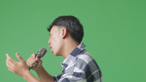 close up side view of young asian teen boy holding a microphone and rapping on the the green screen background