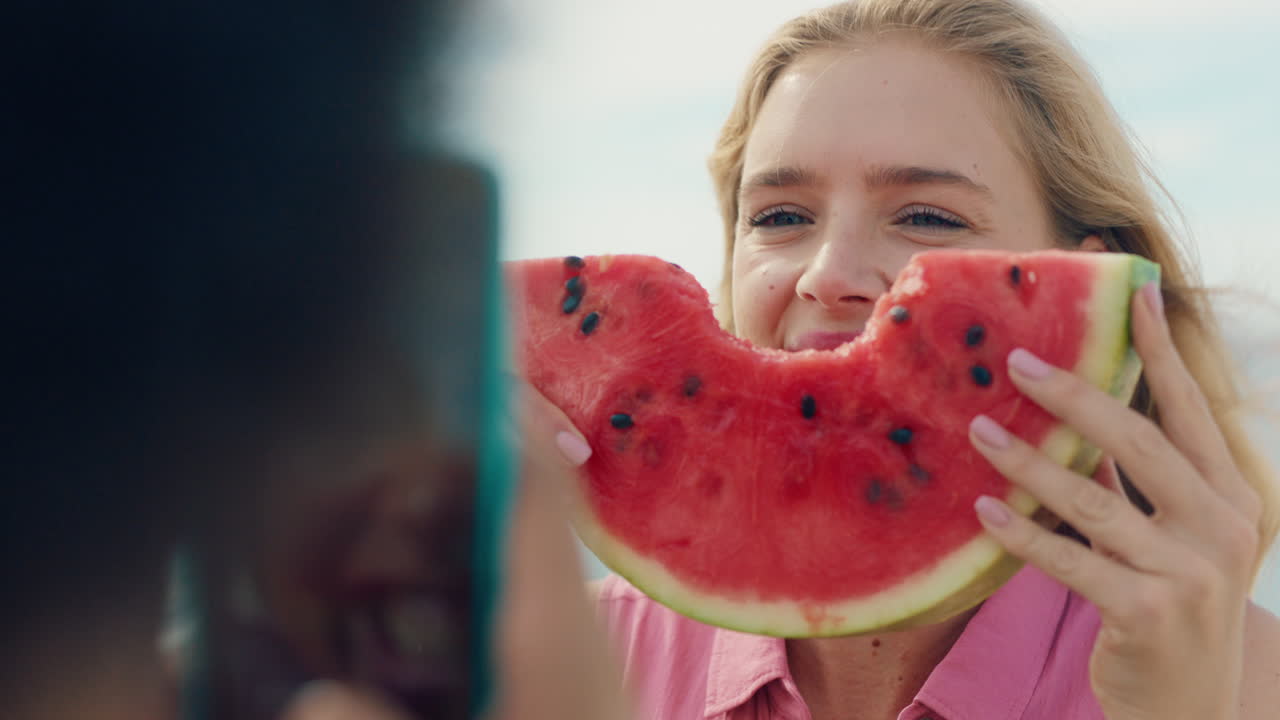 hermosa mujer rubia comiendo sandía en la playa posando para una amiga tomando fotos usando un teléfono inteligente amigas compartiendo el día de verano en las redes sociales divirtiéndose en la playa 4k