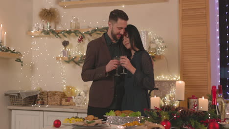 loving couple standing at christmas dinner table, holding champagne glasses and tenderly embracing each other