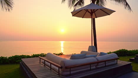 an on an elevated deck and under palm trees and a beach umbrella is an empty cushioned lounger looking out on a colorful ocean sunset