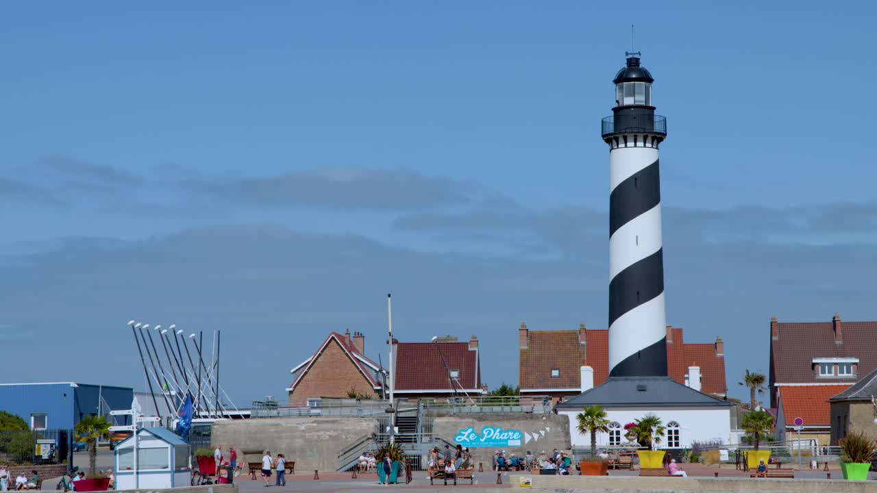 Wide shot of lighthouse, tourists, beach town, bright daylight, static camera, vibrant summer atmosphere