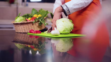 a slow mo side angle close up view of a lady chef cutting cabbage with knife in the kitchen, a basket of veggies with tomatoes and garlic on the table, the chef wore and orange colored apron