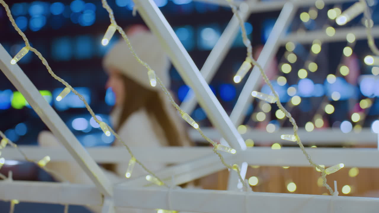 Blurred view of woman in white beanie and winter jacket walking past glowing golden holiday lights, festive atmosphere with decorative string lights and illuminated structures