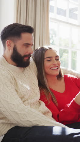 video vertical de una feliz pareja caucásica sentada en el sofá y usando un teléfono inteligente