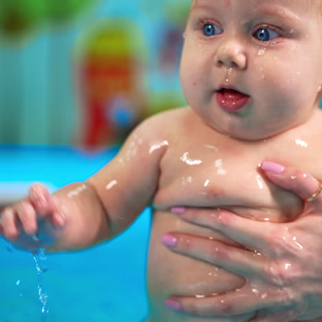 chubby caucasian baby is in the swimming pool. female coach moves the kid in water and he splashes it happily.