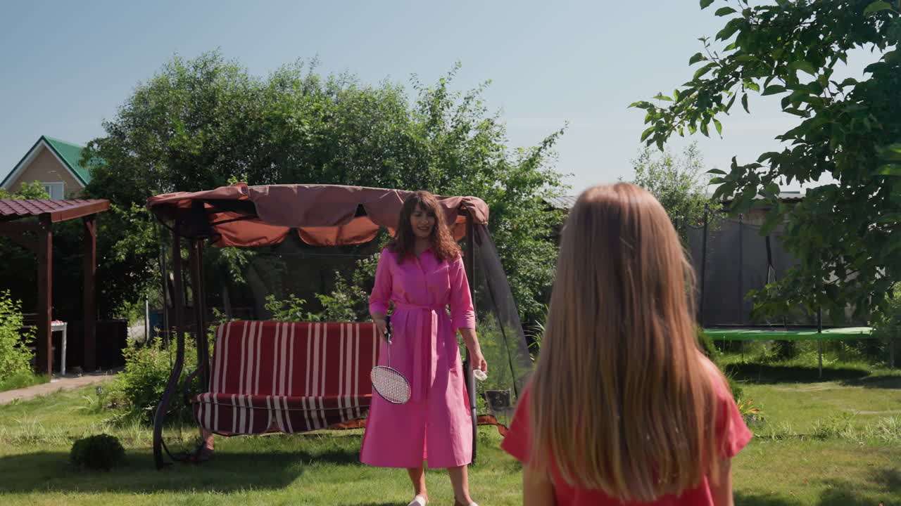 Sunny Backyard White Mother Playing Badminton With Daughter, Lawn Swing And Striped Canopy In Background, Pink Dress Motion, Gentle Coaching And Laughter, Casual Summer Sport Session