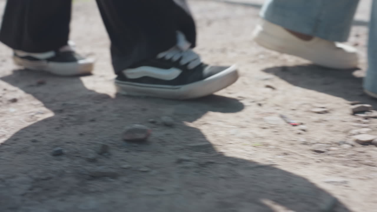 Low angle close up showing sneaker clad feet of group walking along dusty rough rural road in bright sunlight, casting moving shadows across uneven ground