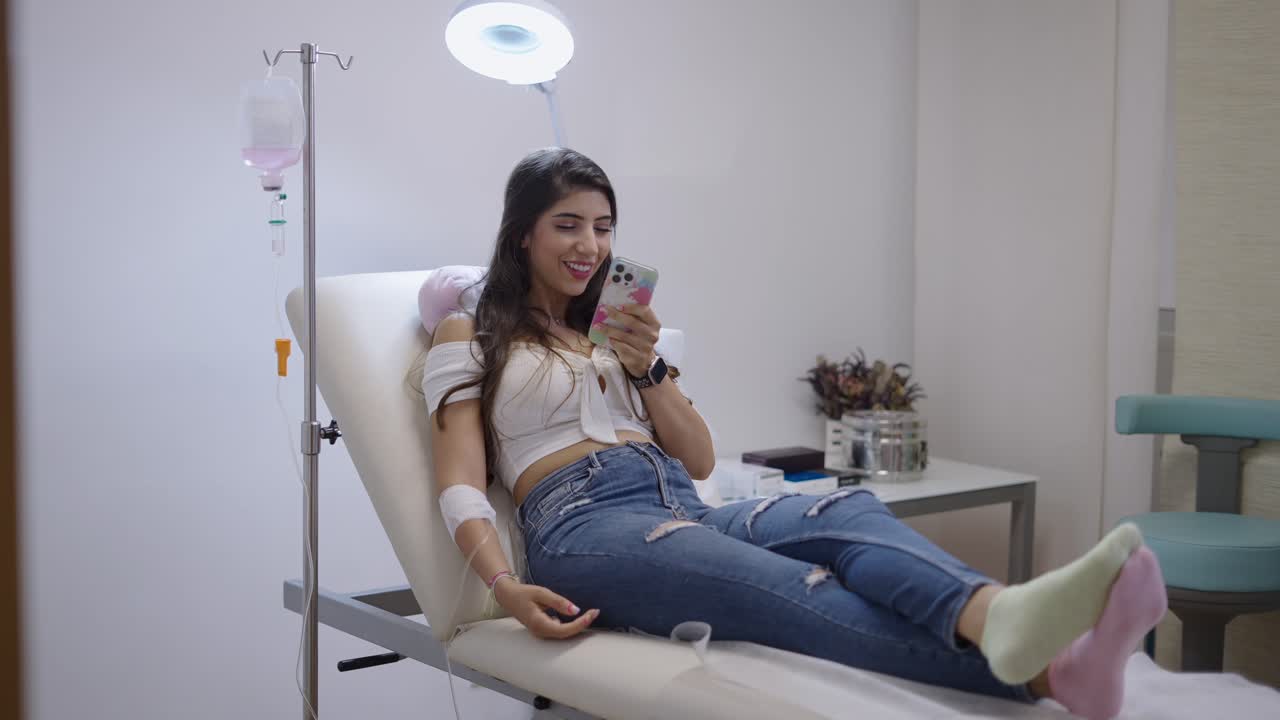 Giovane donna che riceve una flebo mentre usa lo smartphone in una clinica.