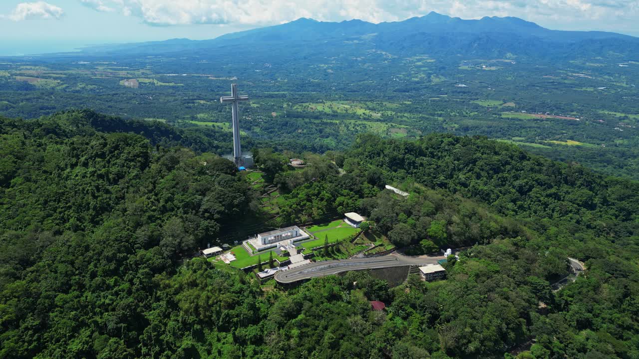 Slow push aerial highlighting the towering Mt. Samat National Shrine, gradually revealing its cross monument framed by lush forested hills and sweeping valleys in Pilar, Bataan, Philippines