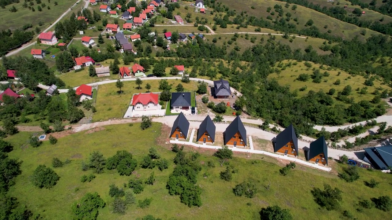 En drone afslører spredte træer på en græsklædt bjergskråning, før en række hytter og røde taghuse dukker op i Zlatibor, Serbien på en klar sommerdag.