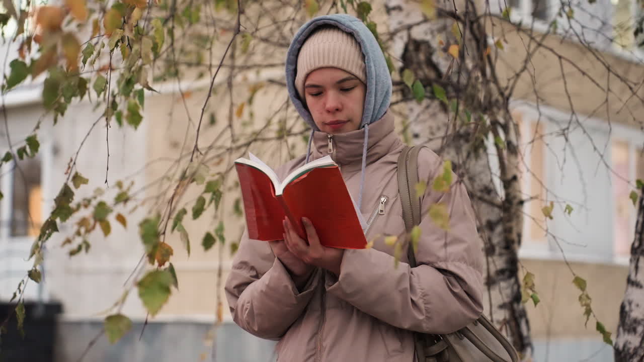 Light skin lady in warm beige coat and hoodie stands quietly under tree with yellow leaves, flipping pages of red book during autumn, with soft focus house and branches forming gentle natural frame