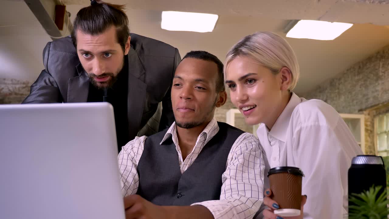 Tre lavoratori multietnici discutono un'idea al portatile, ridendo, concetto di coworking, concetto di comunicazione
