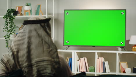 middle eastern man watching tv with chroma key close-up. person watches television with green screen in living room. wearing traditional islamic clothes