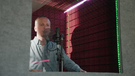 a man wearing a blue long sleeve shirt stands in front of a studio microphone, holding a headphone close to his ear in a red soundproofed room. the scene captures a moment of careful listening