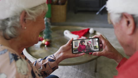 pareja caucásica mayor usando un teléfono inteligente para una videollamada de navidad con una familia sonriente en la pantalla