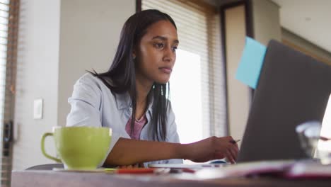 Frau-Mit-Gemischter-Abstammung,-Die-Einen-Laptop-Benutzt-Und-In-Ein-Notizbuch-Schreibt,-Kaffee-Trinkt-Und-Von-Zu-Hause-Aus-Arbeitet