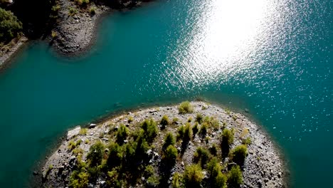 Aerial-flyover-over-islands-with-sunlit-trees-in-the-waters-of-Lac-de-Salanfe-in-Valais,-Switzerland-on-a-sunny-autumn-day-in-the-Swiss-Alps-with-a-pan-up-view-to-surrounding-alpine-peaks