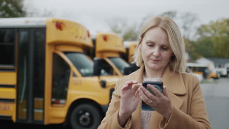 A-teacher-uses-a-smartphone,-stands-in-the-school-yard-against-the-background-of-yellow-school-buses.