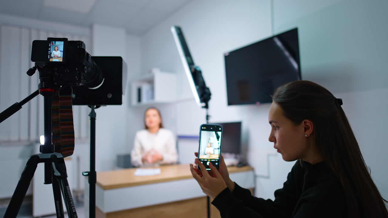 Brunette girl with long hair takes photos on her smartphone. Camera on tripod filming a woman talking at backdrop.