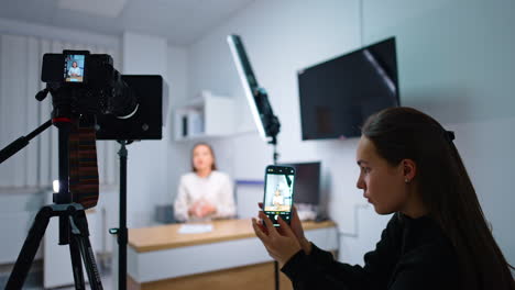 brunette girl with long hair takes photos on her smartphone. camera on tripod filming a woman talking at backdrop.
