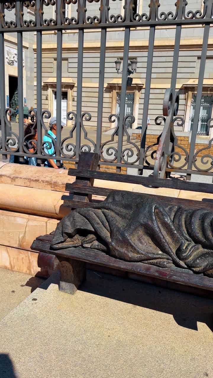 Madrid, Spagna, 2 agosto 2025: Scultura in bronzo di una persona che dorme su una panchina. Vista dettagliata della scultura di Gesù Senzatetto o di una figura in bronzo simile, coperta da una coperta su una panchina di legno.