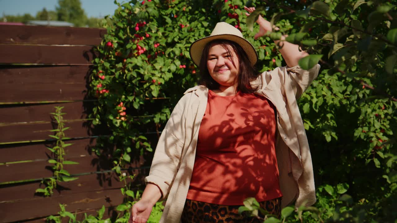 Wanita Tersenyum di Kebun dengan Topi dan Keranjang Apel Menikmati Hari yang Indah Dikelilingi oleh Tumbuhan Hijau Subur dan Pepohonan Berbuah yang Semarak di Surga Halaman Belakangnya