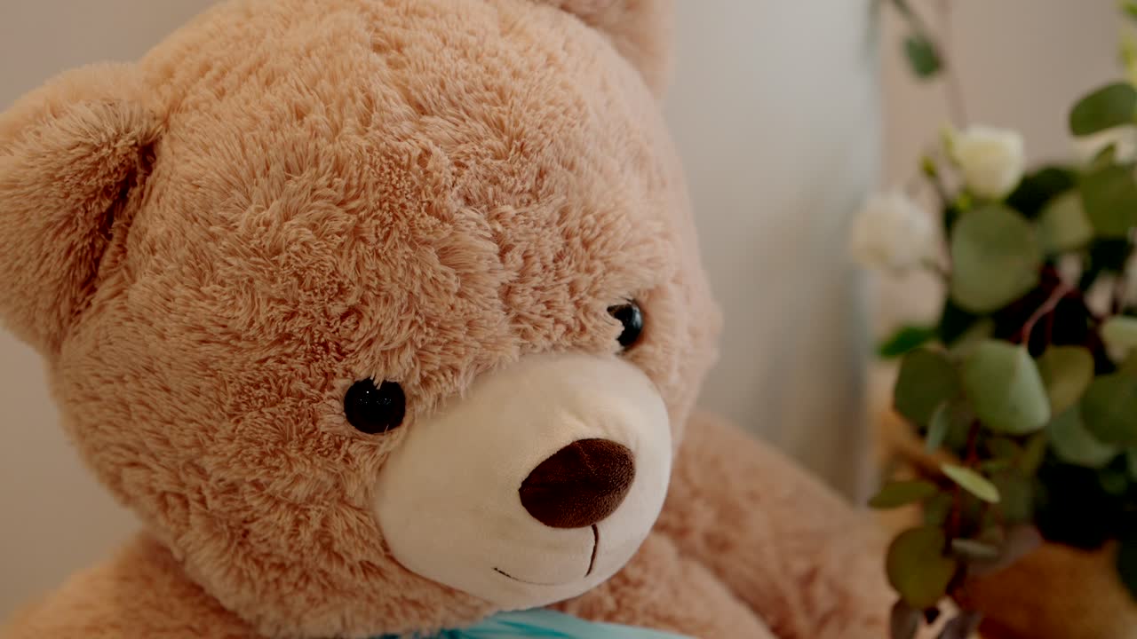 close up of soft teddy bear with blue ribbon and flowers in background indoors