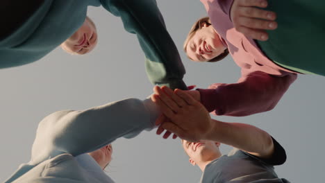 upward view of people in hoodies placing their hands together as team members with sunlight creating a glowing effect around them, capturing team spirit and unity as they smile in outdoor setting